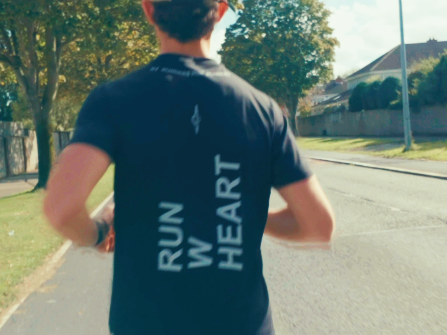 Corredor de espaldas con camiseta negra «RUN WITH HEART» por una calle residencial al sol